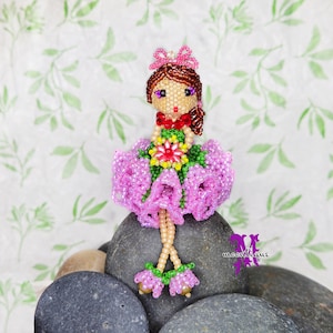 Könnte beinhalten: Eine Perlenpuppe mit rosa und grüner Kleidung, die eine Blume hält und eine rosa Krone trägt, sitzt auf einem Stapel grauer Steine.