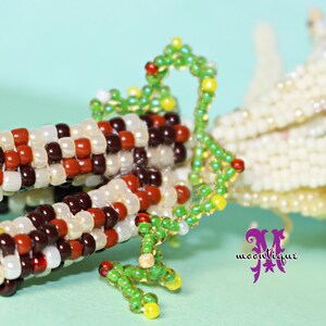 May include: A beaded corn sculpture with kernels in shades of cream, brown, and red. Green beaded vines and yellow accents add detail. The corn's husk is made of cream-colored beads. The sculpture is set against a light blue background.