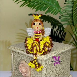 May include: A beaded bee figurine with a yellow and brown skirt, a striped torso, and white wings. The bee has a yellow crown and brown hair. The figurine is sitting on a decorative, off-white box with a rose on the front.
