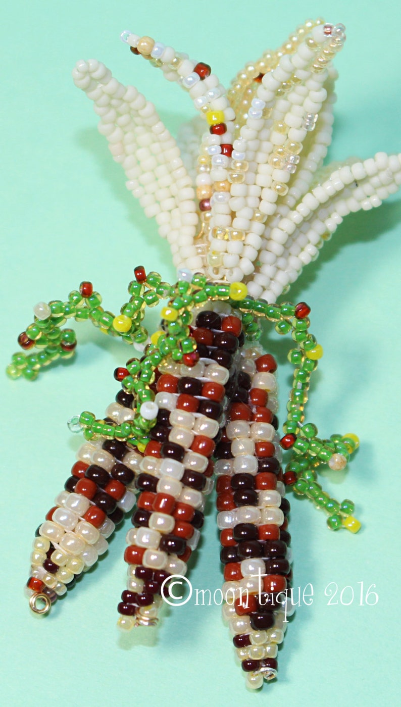 May include: A beaded corn sculpture with three ears of corn in shades of brown, red, and cream. The husks are cream-colored, and green beaded vines are wrapped around the corn. The text "&copy;moontique 2016" is visible.