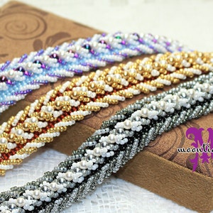 May include: Four beaded bracelets in blue, gold, silver, and white are displayed on a brown box. The bracelets are intricately woven with small beads, creating a textured, braided appearance. The bracelets are a beautiful example of handmade jewellery.