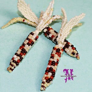 May include: Close-up of two beaded corn on the cob ornaments. The corn kernels are a mix of red, brown, white, and cream-colored beads. The husks are made of cream-colored beads. The ornaments are on a light blue background.