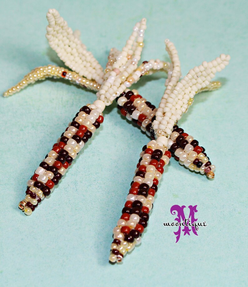 May include: Close-up of two beaded corn on the cob ornaments. The corn kernels are a mix of red, brown, white, and cream-colored beads. The husks are made of cream-colored beads. The ornaments are on a light blue background.