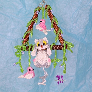 May include: A beaded cat figurine hanging from a beaded branch with two pink beaded birds. The cat is gray and white with yellow eyes. The branch is brown with green vines.