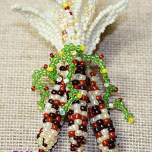 May include: A beaded corn sculpture with three ears of corn in shades of red, brown, cream, and white. The husks are cream-colored and the leaves are green. The sculpture is on a tan surface.