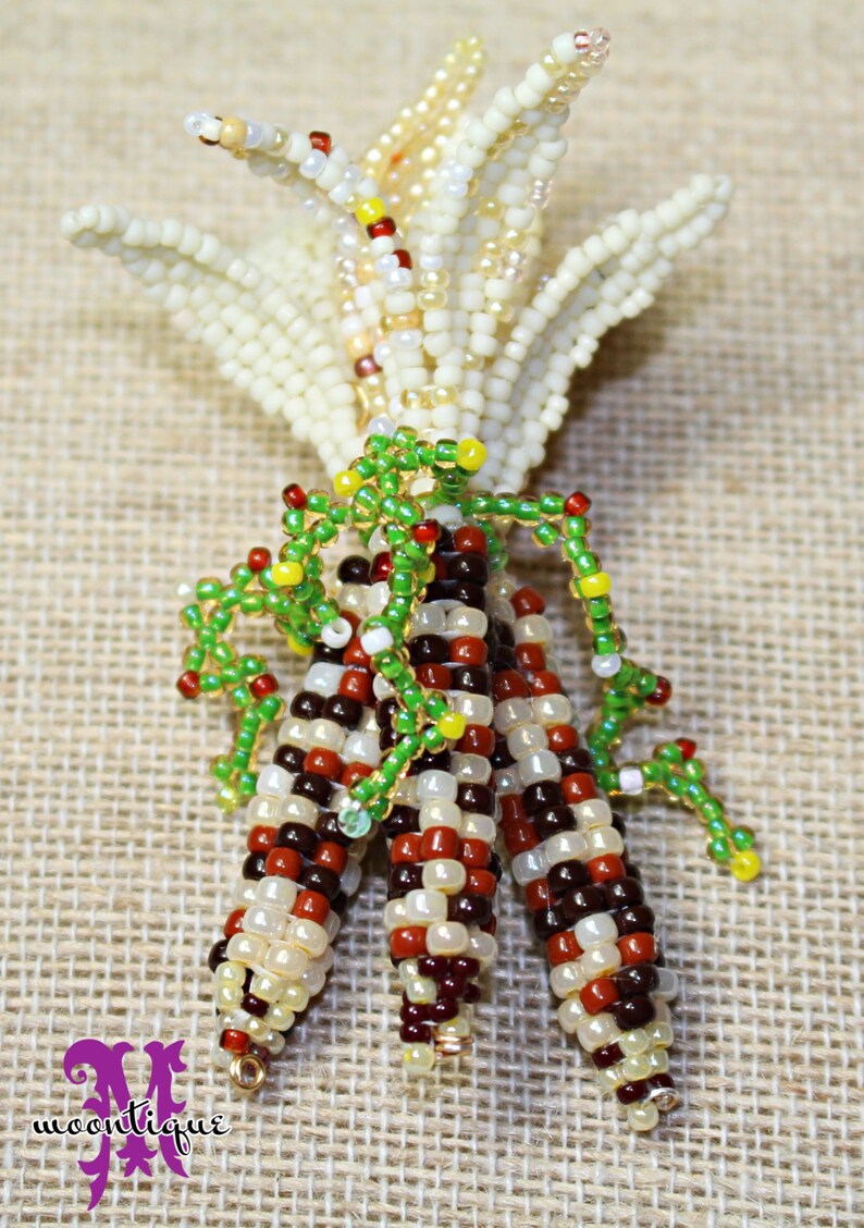 May include: A beaded corn sculpture with three ears of corn in shades of red, brown, cream, and white. The husks are cream-colored and the leaves are green. The sculpture is on a tan surface.