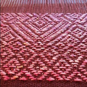 May include: Close-up of a woven fabric with a pink and purple diamond pattern. The fabric has a textured surface and a subtle sheen.