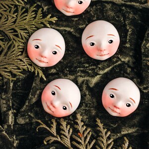 May include: Four white porcelain doll faces with painted features, including rosy cheeks, are arranged on a green velvet background. The faces are slightly tilted and have a whimsical expression.