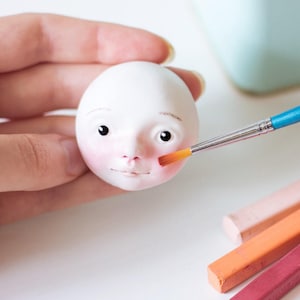May include: A white ceramic doll head with painted features is being painted with a small brush. The doll has brown eyes, a pink nose, and a red mouth. The brush is being used to add a blush to the cheeks. There are four pastel crayons in the background.