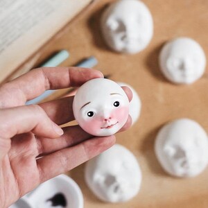 May include: A white ceramic doll head with painted features, including pink cheeks, brown eyes, and a small nose. The doll head is being held in a hand, and there are other unfinished doll heads in the background.