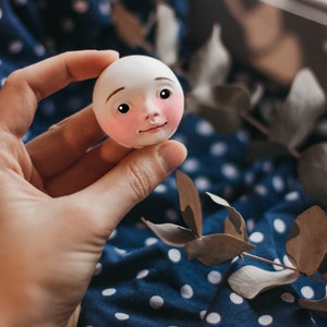 Pode incluir: Uma cabe&ccedil;a de boneca de porcelana branca com tra&ccedil;os pintados, incluindo bochechas rosadas, olhos castanhos e um nariz pequeno. A cabe&ccedil;a da boneca est&aacute; sendo segurada em uma m&atilde;o contra um fundo de tecido azul e branco com bolinhas.