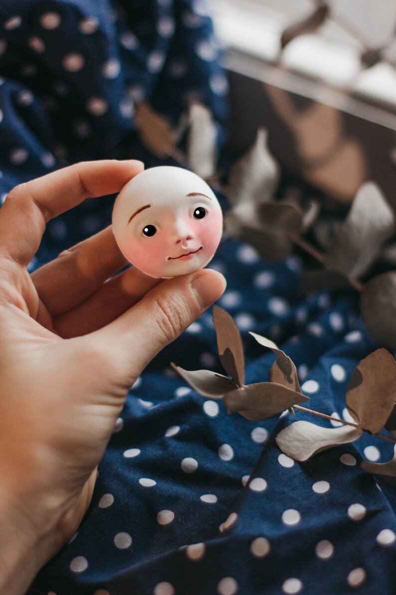 Pode incluir: Uma cabe&ccedil;a de boneca de porcelana branca com tra&ccedil;os pintados, incluindo bochechas rosadas, olhos castanhos e um nariz pequeno. A cabe&ccedil;a da boneca est&aacute; sendo segurada em uma m&atilde;o contra um fundo de tecido azul e branco com bolinhas.