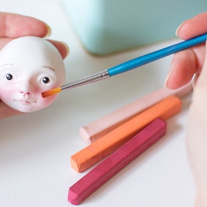 May include: A person paints the face of a small white doll with a blue handled paintbrush. The doll has a pink blush on its cheeks.  Four pastel colored sticks are on the table.