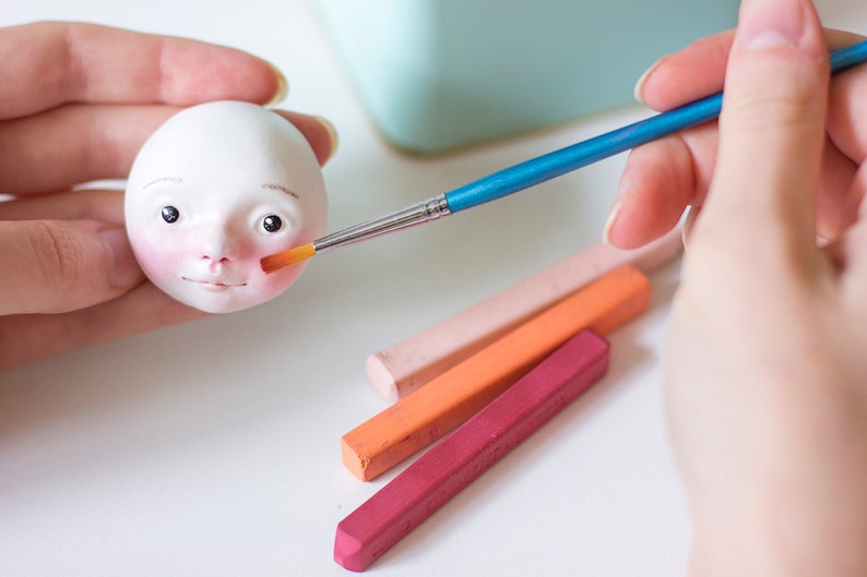 May include: A person paints the face of a small white doll with a blue handled paintbrush. The doll has a pink blush on its cheeks.  Four pastel colored sticks are on the table.
