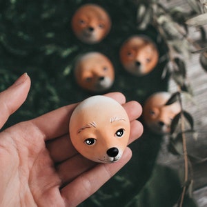 Pode incluir: Uma m&atilde;o segura uma pequena escultura de cer&acirc;mica pintada &agrave; m&atilde;o de uma cabe&ccedil;a de raposa. A escultura &eacute; de cor laranja claro com detalhes brancos e olhos pretos. A escultura &eacute; cercada por outras esculturas de cabe&ccedil;a de raposa.