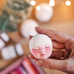 Pode incluir: Uma cabe&ccedil;a de boneca de cer&acirc;mica branca com os olhos fechados e bochechas rosadas. A cabe&ccedil;a da boneca est&aacute; sendo segurada por uma m&atilde;o.