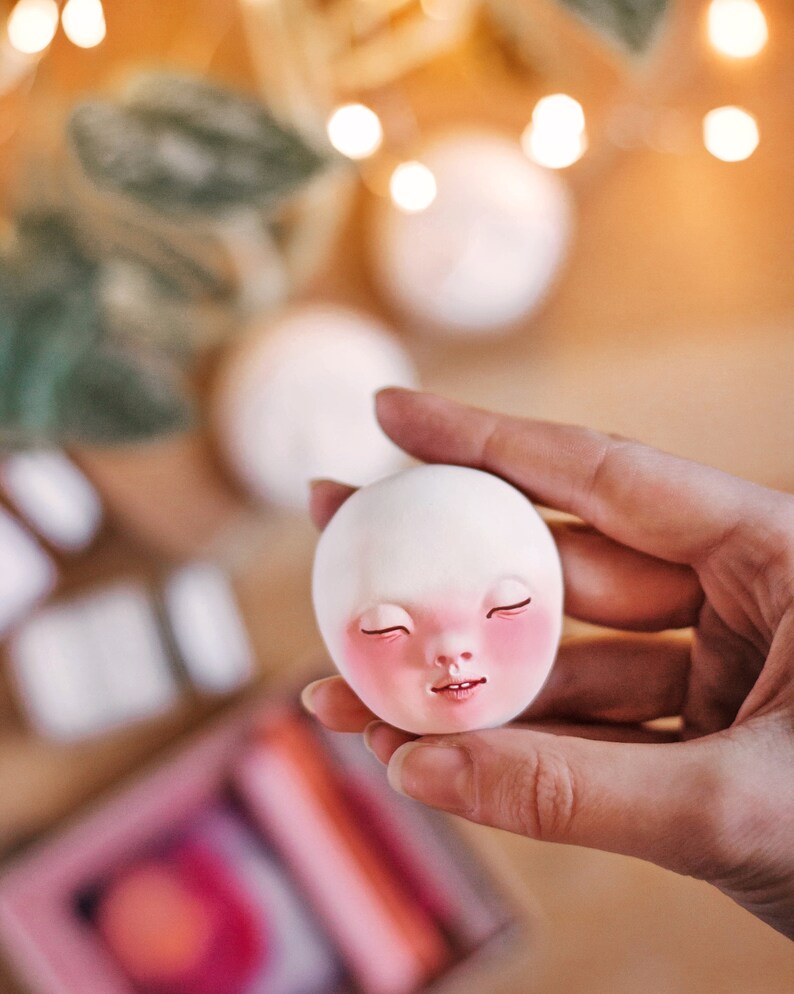 Pode incluir: Uma cabe&ccedil;a de boneca de cer&acirc;mica branca com os olhos fechados e bochechas rosadas. A cabe&ccedil;a da boneca est&aacute; sendo segurada por uma m&atilde;o.