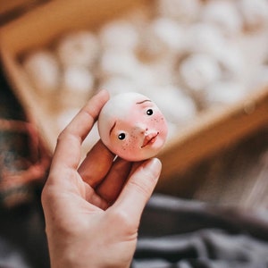 Pode incluir: Uma m&atilde;o segura uma pequena cabe&ccedil;a de boneca branca com tra&ccedil;os pintados, incluindo bochechas rosadas, olhos castanhos e uma boca rosa. A cabe&ccedil;a da boneca &eacute; feita de um material liso, semelhante &agrave; cer&acirc;mica.
