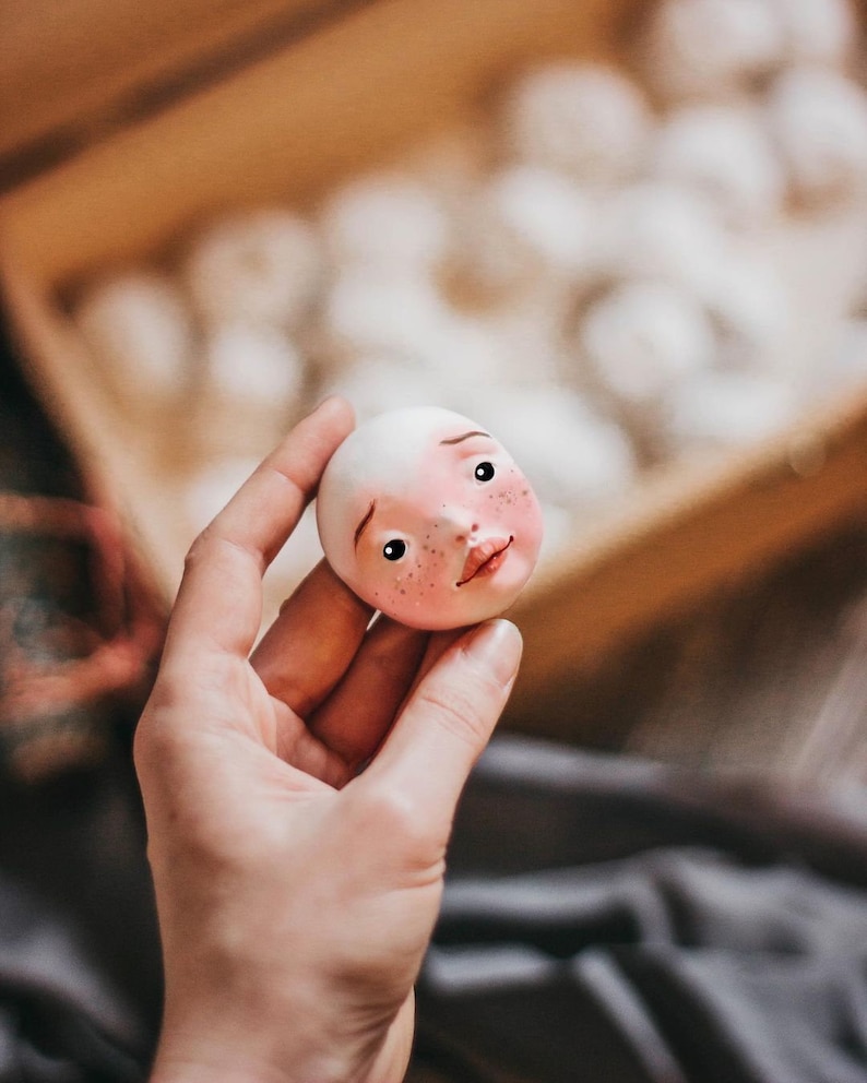 Pode incluir: Uma m&atilde;o segura uma pequena cabe&ccedil;a de boneca branca com tra&ccedil;os pintados, incluindo bochechas rosadas, olhos castanhos e uma boca rosa. A cabe&ccedil;a da boneca &eacute; feita de um material liso, semelhante &agrave; cer&acirc;mica.