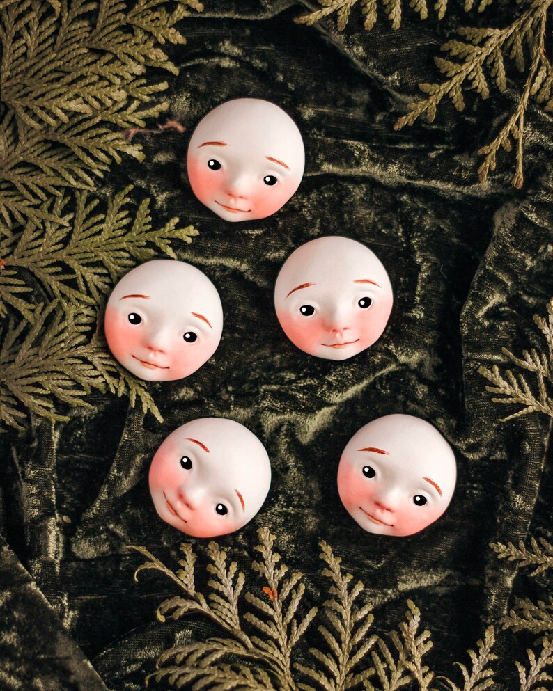 May include: Four white porcelain doll faces with painted features, including rosy cheeks, are arranged on a green velvet background. The faces are slightly tilted and have a whimsical expression.
