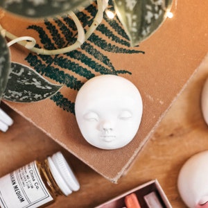 Pode incluir: Escultura de rosto de cer&acirc;mica branca com os olhos fechados e uma pequena boca. A escultura est&aacute; sentada sobre uma superf&iacute;cie marrom com um padr&atilde;o de samambaia verde.