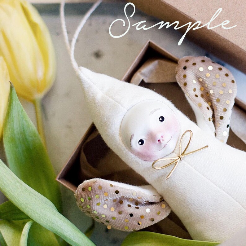 May include: A white plush toy with a human-like face and gold polka dot wings. The toy is in a brown box with the word "Sample" written on it.