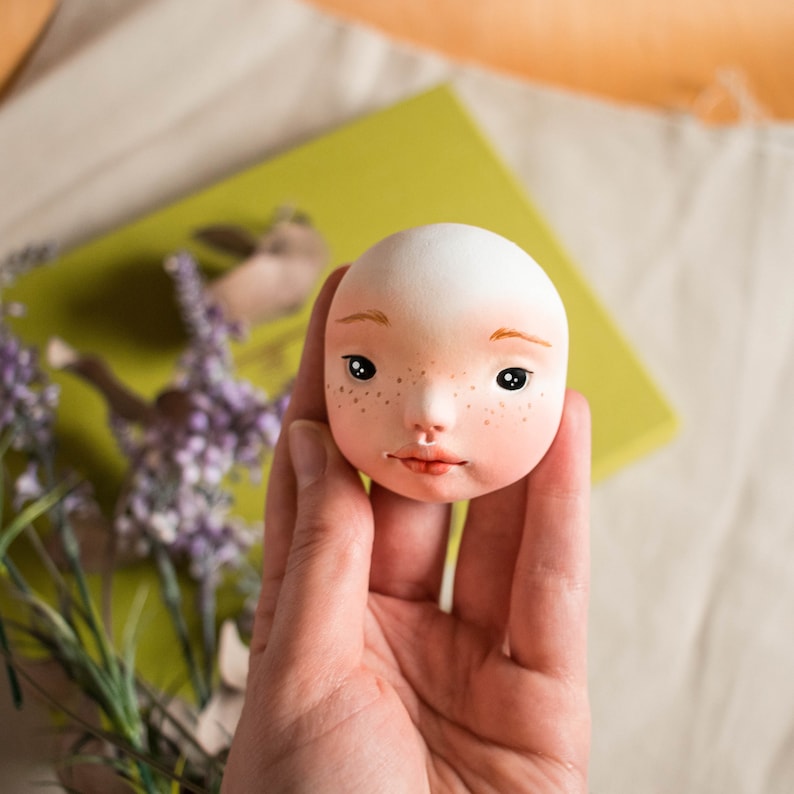 Pode incluir: Uma cabe&ccedil;a de boneca de porcelana branca com olhos castanhos pintados, um nariz rosa e sardas. A boca da boneca est&aacute; ligeiramente aberta, revelando um sorriso rosa pintado.