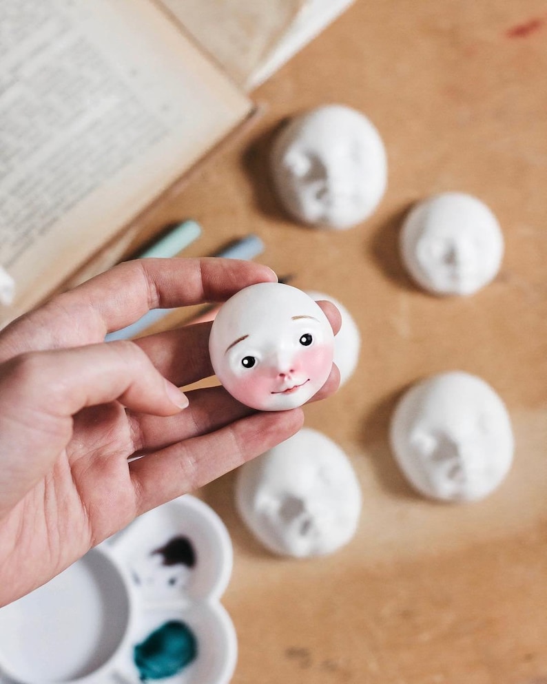 May include: A white ceramic doll head with painted features, including pink cheeks, brown eyes, and a small nose. The doll head is being held in a hand, and there are other unfinished doll heads in the background.