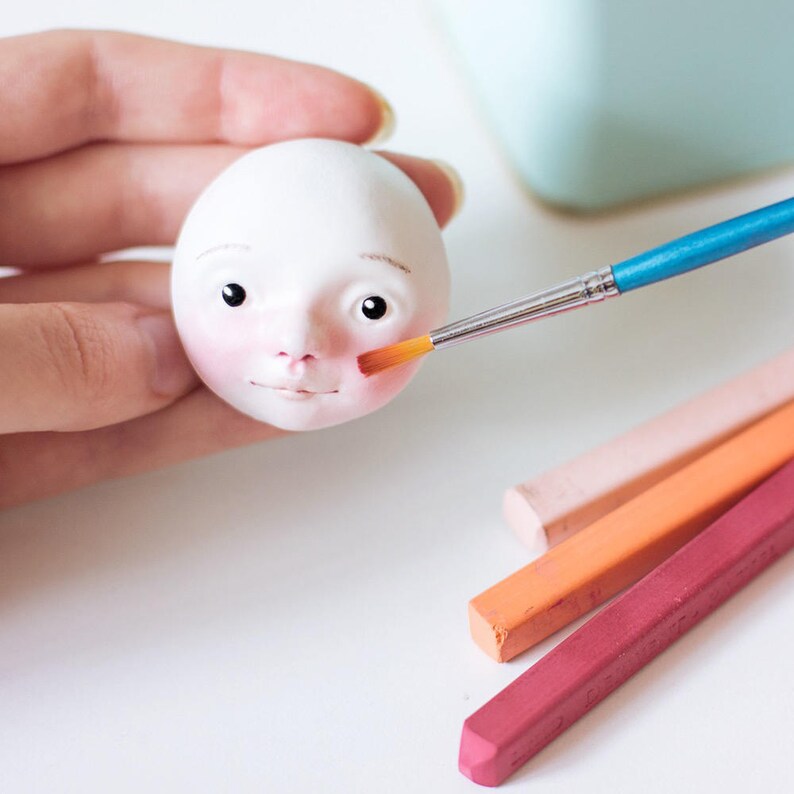 May include: A white ceramic doll head with painted features is being painted with a small brush. The doll has brown eyes, a pink nose, and a red mouth. The brush is being used to add a blush to the cheeks. There are four pastel crayons in the background.