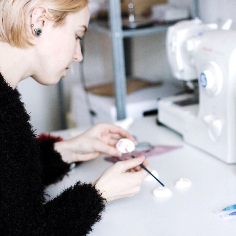 May include: A person is painting small white ceramic hearts with a black paintbrush. The hearts are on a white surface. There is a sewing machine in the background.