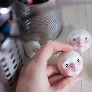 May include: Two porcelain doll heads with painted features, one in a hand and one on a table. The doll heads have pink cheeks, closed mouths, and blue eyes.