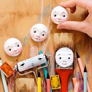 May include: A wooden surface with various art supplies, including paintbrushes, paint rollers, pencils, and watercolor paints. There are also four white ceramic faces with painted features, some of which are being held by a hand.