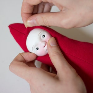 May include: A hand is holding a red fabric doll with a white face and painted eyes. The doll is wearing a red hooded garment.