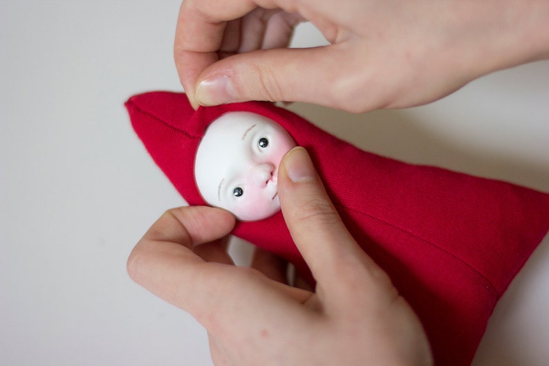 May include: A hand is holding a red fabric doll with a white face and painted eyes. The doll is wearing a red hooded garment.