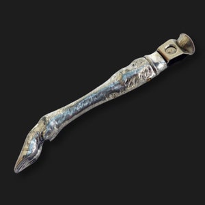 May include: A silver metal cigar cutter shaped like a leg with a textured surface. The cutter has a small hole in the top for cutting cigars.