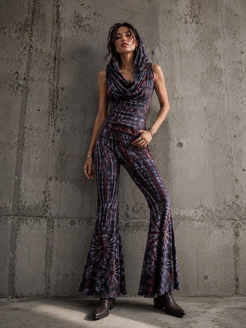 K&ouml;nnte beinhalten: Ein &auml;rmelloser Jumpsuit mit Kapuze, Wasserfall-Ausschnitt und ausgestellten Hosenbeinen. Das Kleidungsst&uuml;ck besteht aus einem dunklen Stoff mit einem Batikmuster in Schwarz-, Grau- und Dunkelrott&ouml;nen, akzentuiert durch kreisf&ouml;rmige Motive.