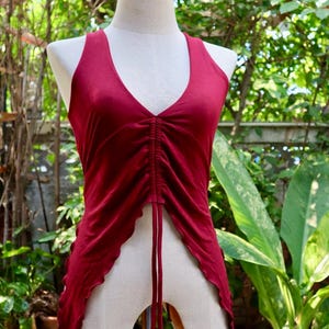Könnte beinhalten: Burgunderfarbenes Neckholder-Top mit V-Ausschnitt und gerafften Details vorne. Das Top hat einen fließenden, asymmetrischen Saum und wird auf einer Schaufensterpuppe präsentiert. Der Hintergrund zeigt üppiges grünes Laub.