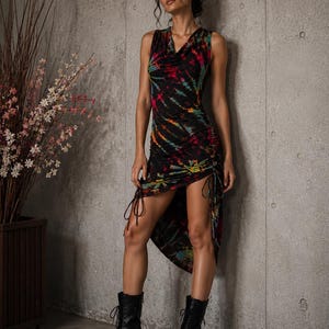 May include: A sleeveless tie-dye dress in black, red, and teal, with a draped neckline and ruched detailing. The dress is worn with black combat boots. The model is standing next to a concrete wall.