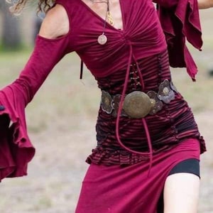 Pode incluir: Um vestido comprido de cor bordô com um design de ombro descoberto e uma bainha com babados. O vestido tem um corpete justo com um detalhe de laço e uma saia fluida. O vestido é adornado com um cinto com uma fivela circular grande e vários colares.