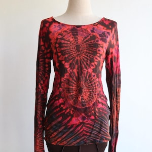 May include: A long-sleeved tie-dye shirt in shades of red, black, and purple. The shirt features a round neckline and a central sunburst design. The fabric appears lightweight and the sleeves are fitted.