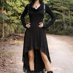 May include: A black dress with a high-low hemline and lace trim. The long-sleeved dress features a cowl neckline and is paired with black combat boots. A black harness is worn over the dress. The model is standing on a dirt path in a wooded area.