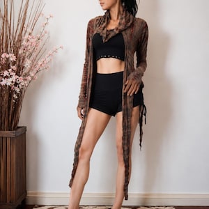 Könnte beinhalten: Eine Frau trägt eine braun-rote Batik-Strickjacke mit langen Ärmeln und einem drapierten Kragen. Sie trägt auch ein schwarzes Crop-Top und schwarze Shorts. Das Outfit wird mit einem Teppich mit Tigerstreifenmuster kombiniert.