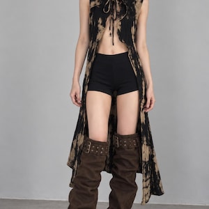 May include: A sleeveless, hooded long vest featuring a black and tan tie-dye pattern with a front tie closure. The outfit includes solid black shorts and dark brown, knee-high, slouchy boots with buckle strap details at the top.