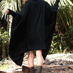 Op de afbeelding: Een zwarte, oversized poncho-stijl trui met een col en lange mouwen. De trui wordt gecombineerd met bruine laarzen en een zwarte hoed. De achtergrond heeft weelderig groen gebladerte.