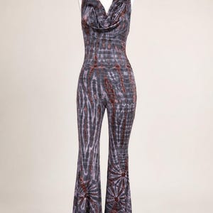 May include: A sleeveless jumpsuit featuring a cowl neckline and an attached hood. The garment has a fitted bodice and flared pant legs, decorated with a dark gray and rust-colored tie-dye pattern that creates a radial design at the bottom hems.