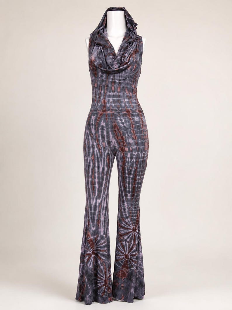May include: A sleeveless jumpsuit featuring a cowl neckline and an attached hood. The garment has a fitted bodice and flared pant legs, decorated with a dark gray and rust-colored tie-dye pattern that creates a radial design at the bottom hems.