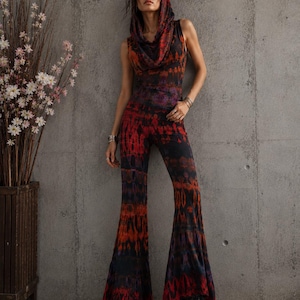 K&ouml;nnte beinhalten: Ein &auml;rmelloser Jumpsuit mit Kapuze, Wasserfallkragen und ausgestellten Hosenbeinen. Das Kleidungsst&uuml;ck besteht aus einem Stoff mit Batikmuster in den Farben Schwarz, Dunkelrot und Orange. Der Jumpsuit wird mit schwarzen Schn&uuml;rstiefeln kombiniert.