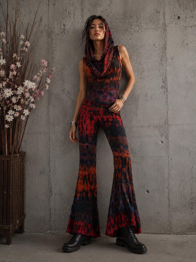K&ouml;nnte beinhalten: Ein &auml;rmelloser Jumpsuit mit Kapuze, Wasserfallkragen und ausgestellten Hosenbeinen. Das Kleidungsst&uuml;ck besteht aus einem Stoff mit Batikmuster in den Farben Schwarz, Dunkelrot und Orange. Der Jumpsuit wird mit schwarzen Schn&uuml;rstiefeln kombiniert.