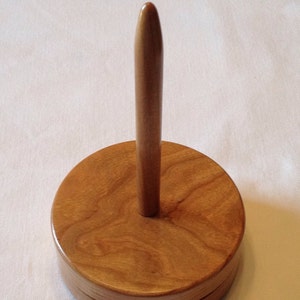 May include: A wooden yarn swift with a tall, slender spindle and a round base. The wood has a natural, light brown color with a grain pattern.