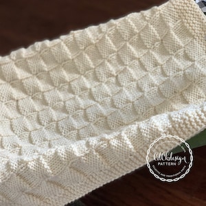 May include: Cream-coloured knitted blanket with a textured diamond pattern, resting in a vintage green wagon. The blanket's edges have a ribbed border. A black and white checkered ribbon is tied to the wagon. The blanket is a cosy, handmade item.